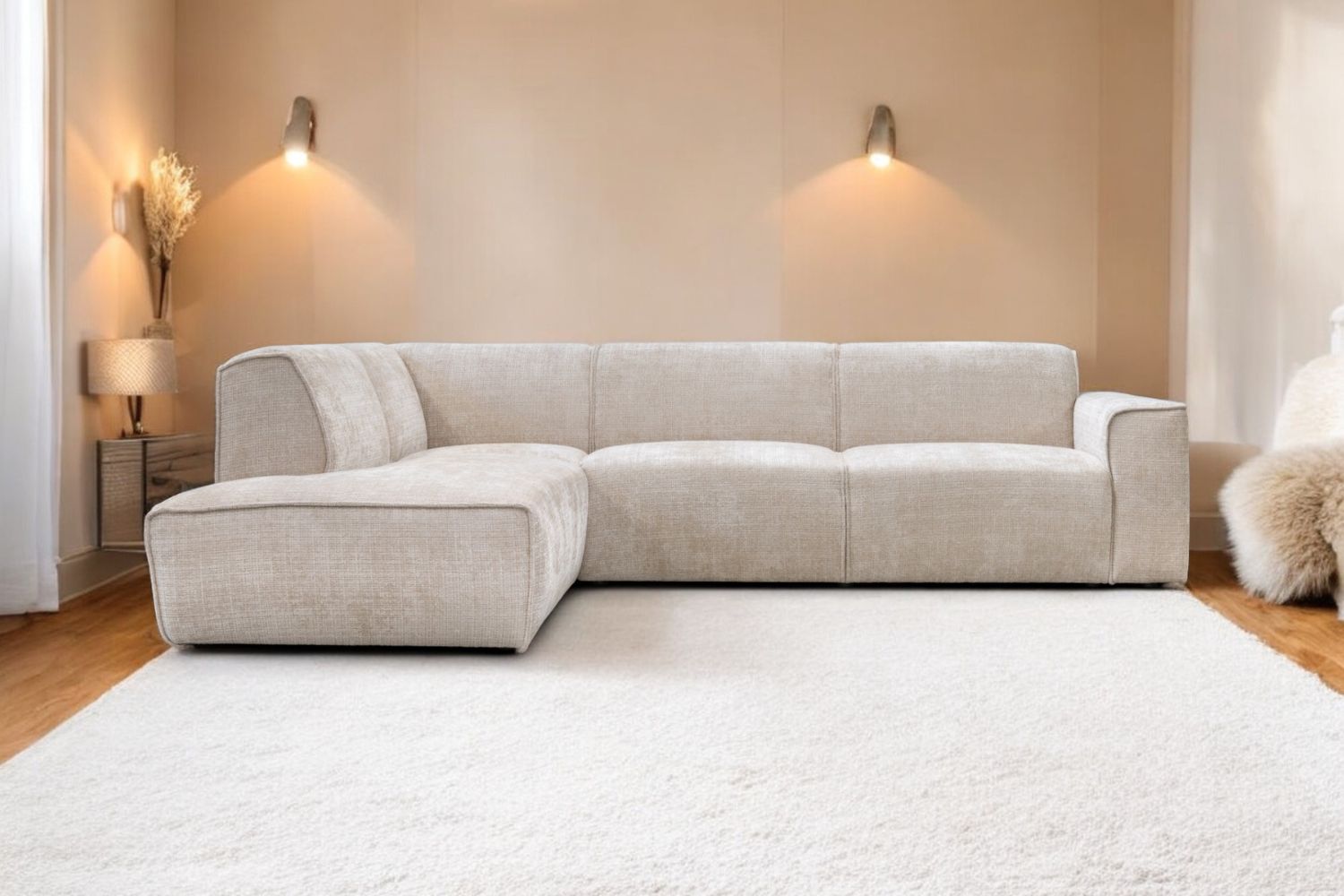 Canapé d'angle beige 4 places dans une pièce éclairé par des spots sur le mur du fond, sur un tapis blanc. 